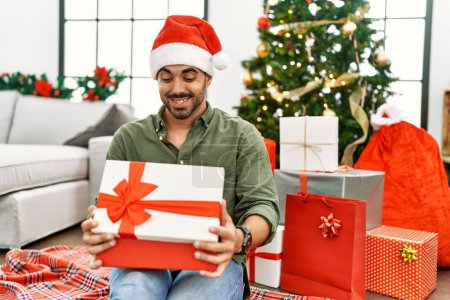 Téléchargez les photos : Jeune homme hispanique cadeau de déballage assis sur le sol par l'arbre de Noël à la maison - en image libre de droit