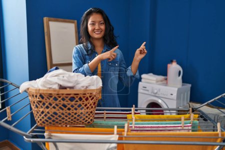 Photo for Young asian woman hanging clothes at clothesline smiling and looking at the camera pointing with two hands and fingers to the side. - Royalty Free Image