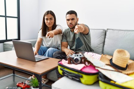 Photo for Young interracial couple packing summer clothes in suitcase looking at laptop pointing with finger to the camera and to you, confident gesture looking serious - Royalty Free Image