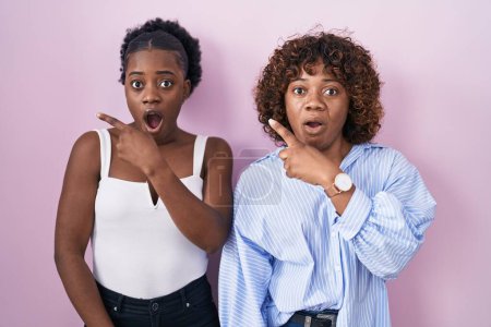 Téléchargez les photos : Two african women standing over pink background surprised pointing with finger to the side, open mouth amazed expression. - en image libre de droit