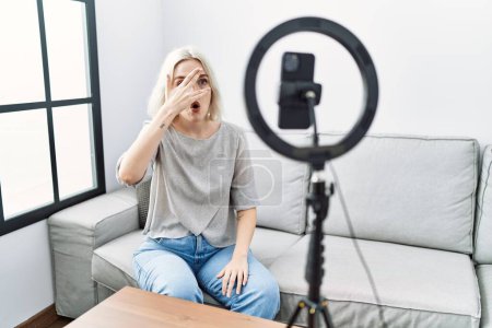 Foto de Young caucasian woman recording vlog tutorial with smartphone at home peeking in shock covering face and eyes with hand, looking through fingers with embarrassed expression. - Imagen libre de derechos