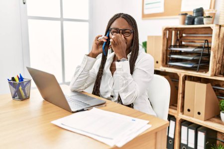 Téléchargez les photos : Femme noire avec des tresses travaillant au bureau parlant au téléphone sentant quelque chose de puant et dégoûtant, odeur intolérable, retenant la respiration avec les doigts sur le nez. mauvaise odeur - en image libre de droit