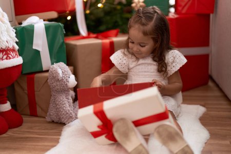 Foto de Adorable chica hispana desempacando regalo sentado en el suelo por árbol de Navidad en casa - Imagen libre de derechos