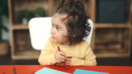 Téléchargez les photos : Adorable étudiante hispanique assise sur la table dessinant sur papier à la maternelle - en image libre de droit