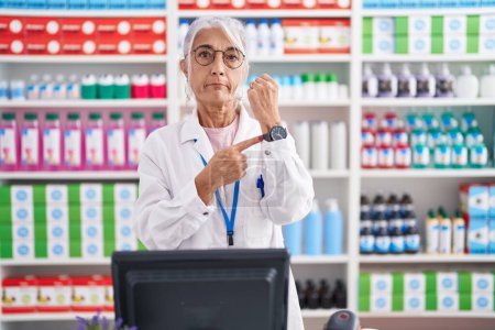 Téléchargez les photos : Femme d'âge moyen avec des tatouages travaillant à la pharmacie pharmacie à la hâte pointant vers regarder le temps, l'impatience, regarder la caméra avec une expression détendue - en image libre de droit