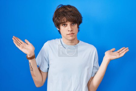 Photo for Hispanic young man standing over blue background clueless and confused expression with arms and hands raised. doubt concept. - Royalty Free Image