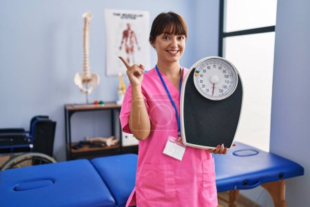 Photo for Young brunette woman as nutritionist holding weighing machine smiling happy pointing with hand and finger to the side - Royalty Free Image