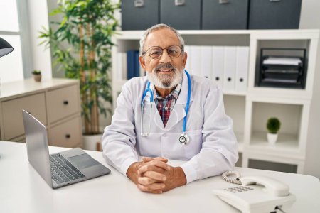 Téléchargez les photos : Homme âgé aux cheveux gris médecin souriant confiant assis sur la table à la clinique - en image libre de droit