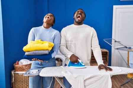 Foto de Joven pareja afroamericana planchando ropa en la lavandería enojada y loca gritando frustrada y furiosa, gritando con ira mirando hacia arriba. - Imagen libre de derechos