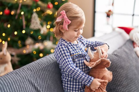 Photo for Adorable hispanic girl playing with toy standing on sofa by christmas tree at home - Royalty Free Image