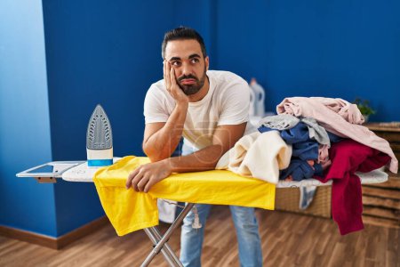 Téléchargez les photos : Young hispanic man tired leaning on ironing board at laundry room - en image libre de droit