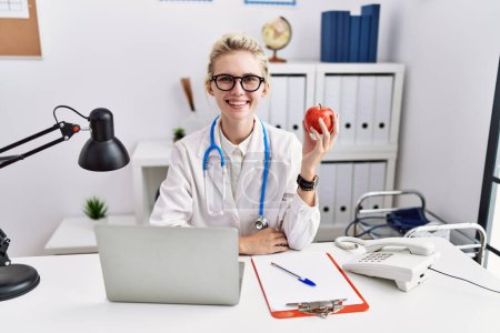 Téléchargez les photos : Jeune femme médecin travaillant à la clinique diététiste regardant positif et heureux debout et souriant avec un sourire confiant montrant les dents - en image libre de droit