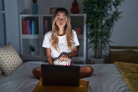 Young blonde woman sitting on the bed at home watching a movie from laptop puffing cheeks with funny face. mouth inflated with air, catching air. 