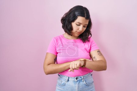 Téléchargez les photos : Jeune femme hispanique debout sur fond rose vérifier l'heure sur la montre-bracelet, détendu et confiant - en image libre de droit