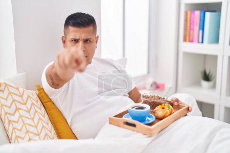 Téléchargez les photos : Jeune homme hispanique manger le petit déjeuner dans le lit pointant du doigt à la caméra et à vous, geste confiant regardant sérieux - en image libre de droit