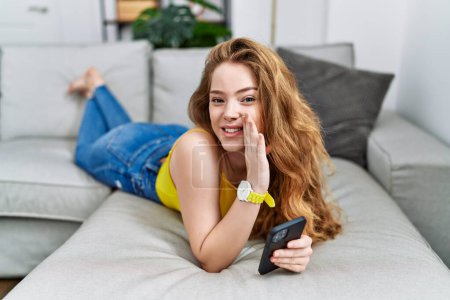 Téléchargez les photos : Jeune femme caucasienne couchée sur le canapé en utilisant smartphone main sur bouche racontant rumeur secrète, chuchotant conversation malicieuse - en image libre de droit