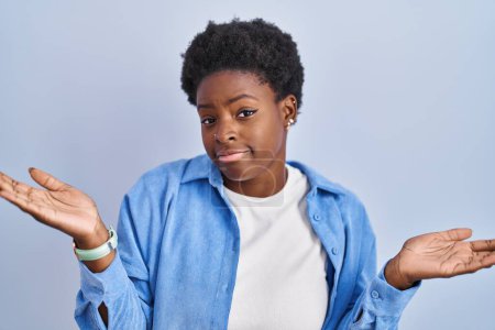 Foto de Mujer afroamericana de pie sobre fondo azul expresión despistada y confusa con los brazos y las manos levantadas. concepto de duda. - Imagen libre de derechos