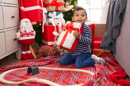 Foto de Adorable niño hispano sosteniendo regalo sentado en el suelo por el árbol de Navidad en casa - Imagen libre de derechos