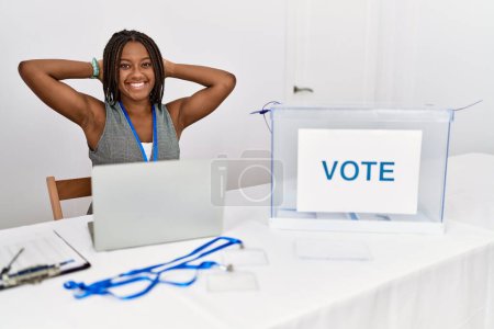 Foto de Mujer afroamericana joven que trabaja en las elecciones políticas sentada por votación relajándose y estirándose, brazos y manos detrás de la cabeza y el cuello sonriendo feliz - Imagen libre de derechos
