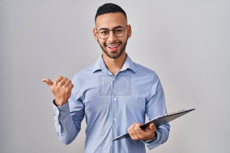 Photo for Young hispanic man wearing business clothes holding clipboard pointing thumb up to the side smiling happy with open mouth - Royalty Free Image