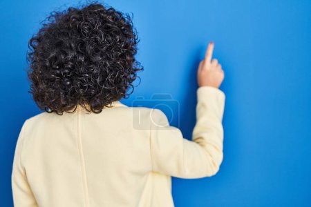 Photo for Young brunette woman with curly hair standing over blue background posing backwards pointing ahead with finger hand - Royalty Free Image