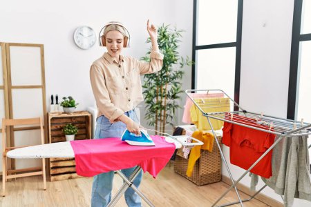 Foto de Young caucasian woman listening to music ironing clothes at laundry room - Imagen libre de derechos