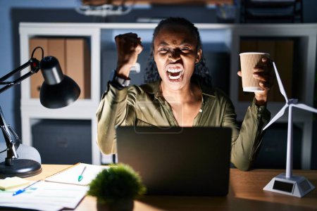 Téléchargez les photos : Femme africaine travaillant à l'aide d'un ordinateur portable la nuit en colère et folle levant le poing frustré et furieux tout en criant de colère. rage et concept agressif. - en image libre de droit