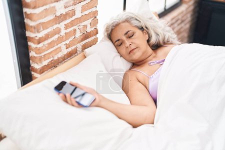 Téléchargez les photos : Moyen-âge femme aux cheveux gris tenant smartphone couché sur le lit dormant dans la chambre - en image libre de droit