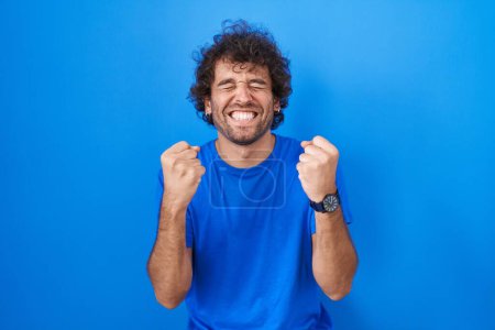 Photo for Hispanic young man standing over blue background excited for success with arms raised and eyes closed celebrating victory smiling. winner concept. - Royalty Free Image