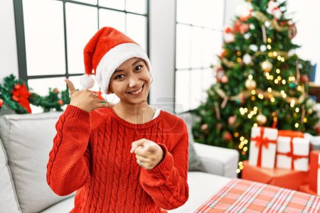Foto de Young hispanic woman with short hair wearing christmas hat sitting on the sofa smiling doing talking on the telephone gesture and pointing to you. call me. - Imagen libre de derechos