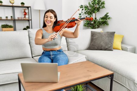 Photo for Young beautiful hispanic woman having online violin class sitting on sofa at home - Royalty Free Image