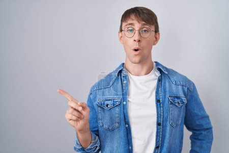 Photo for Caucasian blond man standing wearing glasses surprised pointing with finger to the side, open mouth amazed expression. - Royalty Free Image