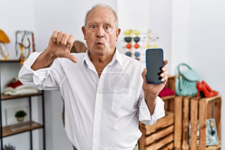 Téléchargez les photos : Homme âgé tenant smartphone au magasin de détail avec le visage en colère, signe négatif montrant aversion avec les pouces vers le bas, concept de rejet - en image libre de droit