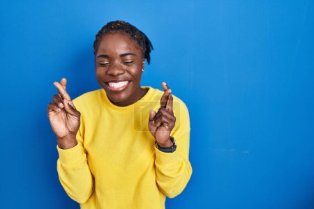 Téléchargez les photos : Beautiful black woman standing over blue background gesturing finger crossed smiling with hope and eyes closed. luck and superstitious concept. - en image libre de droit