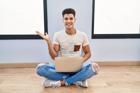 Téléchargez les photos : Jeune homme hispanique utilisant un ordinateur portable à la maison souriant présentation joyeuse et pointant avec la paume de la main en regardant la caméra. - en image libre de droit