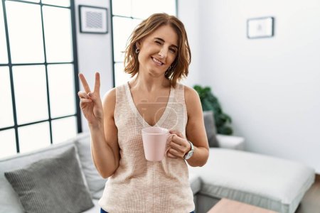 Téléchargez les photos : Femme d'âge moyen buvant une tasse de café à la maison souriant avec le visage heureux clin d'oeil à la caméra faisant signe de victoire avec les doigts. numéro deux. - en image libre de droit