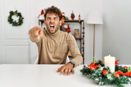 Photo for Young handsome man with beard sitting on the table by christmas decoration pointing displeased and frustrated to the camera, angry and furious with you - Royalty Free Image