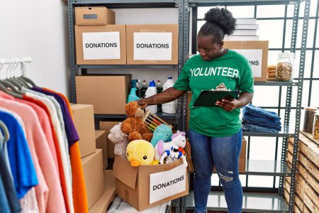 Téléchargez les photos : Jeune femme afro-américaine portant un uniforme bénévole écrivant sur le presse-papiers au centre de charité - en image libre de droit