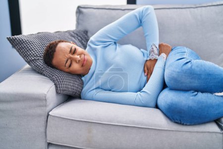 Foto de Mujer afroamericana que sufre de dolor menstrual tumbada en un sofá en casa - Imagen libre de derechos
