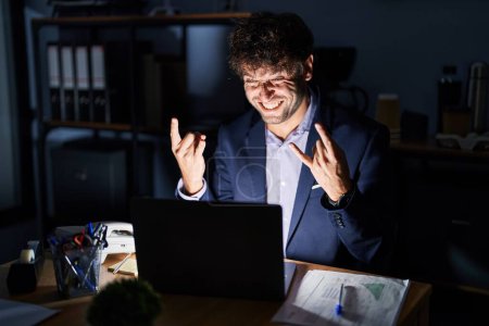 Photo for Hispanic young man working at the office at night shouting with crazy expression doing rock symbol with hands up. music star. heavy concept. - Royalty Free Image