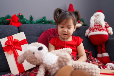 Foto de Adorable chica china sosteniendo oso de peluche sentado en el sofá por la decoración de Navidad en casa - Imagen libre de derechos