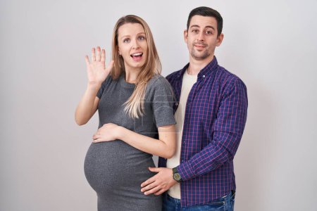 Téléchargez les photos : Jeune couple attendant un bébé debout sur fond blanc renonçant à dire bonjour heureux et souriant, geste de bienvenue amical - en image libre de droit