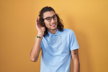 Photo for Young hispanic man standing over yellow background smiling with hand over ear listening an hearing to rumor or gossip. deafness concept. - Royalty Free Image
