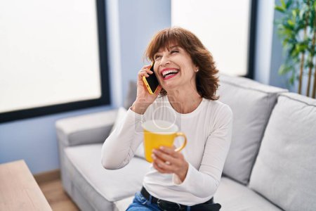Téléchargez les photos : Middle age woman talking on smartphone drinking coffee at home - en image libre de droit