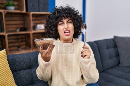 Photo for Young brunette woman with curly hair eating healthy whole grain cereals with spoon clueless and confused expression. doubt concept. - Royalty Free Image