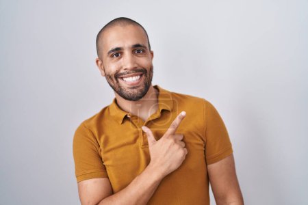Téléchargez les photos : Homme hispanique avec la barbe debout sur fond blanc gai avec un sourire sur le visage pointant avec la main et le doigt vers le côté avec une expression heureuse et naturelle - en image libre de droit
