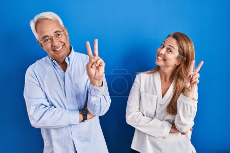 Téléchargez les photos : Couple hispanique du Moyen Age debout sur fond bleu souriant regardant la caméra montrant des doigts faisant signe de victoire. numéro deux. - en image libre de droit