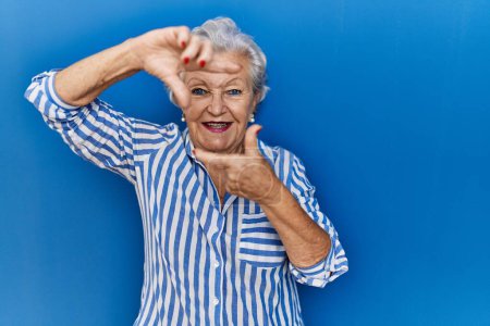 Foto de Mujer mayor con el pelo gris de pie sobre fondo azul sonriendo haciendo marco con las manos y los dedos con la cara feliz. concepto de creatividad y fotografía. - Imagen libre de derechos