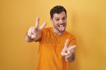 Foto de Joven hombre hispano de pie sobre fondo amarillo sonriendo con la lengua hacia fuera mostrando los dedos de ambas manos haciendo signo de victoria. número dos. - Imagen libre de derechos