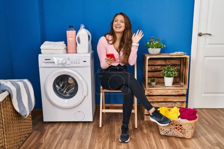 Photo for Young hispanic woman sitting waiting for laundry using smartphone waiving saying hello happy and smiling, friendly welcome gesture - Royalty Free Image